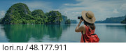 A young woman with a camera admires the serene beauty of Halong Bay surrounded by lush greenery. Стоковое AI-изображение, AI-художник Данила Большаков / Фотобанк Лори