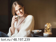 Красивая счастливая молодая женщина пьет кофе. Привлекательная девушка в белом вязаном кардигане возле столика в уютном кафе или дома с чашкой капучино в руках. Мягкий фокус. Стоковое фото, фотограф Андрей Степанов / Фотобанк Лори
