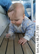 A curious baby is crawling on a wooden floor, fascinated by a clear cup as sunlight filters through the boat's cabin. Стоковое фото, фотограф Данила Большаков / Фотобанк Лори