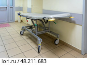 A vacant hospital gurney stands in a well-lit corridor, ready for patient transport during daytime hours in a medical facility. Стоковое фото, фотограф Данила Большаков / Фотобанк Лори