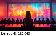 A woman sits in front of a large colorful light display, captivated by the vivid colors as others watch quietly in a contemporary conference setting. Стоковое AI-изображение, AI-художник Данила Большаков / Фотобанк Лори
