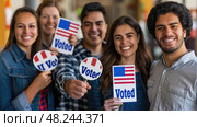 Four young adults stand together smiling, holding Voted signs, celebrating their civic duty. Стоковое AI-изображение, AI-художник Данила Большаков / Фотобанк Лори