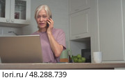 Elderly woman multitasking with phone and laptop in modern kitchen setting. Стоковое видео, видеограф Владимир Федоров / Фотобанк Лори