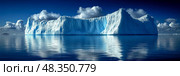 A gigantic iceberg rests in tranquil waters, reflecting shades of blue while clouds drift across the clear sky in a remote Arctic area. Стоковое AI-изображение, AI-художник Данила Большаков / Фотобанк Лори