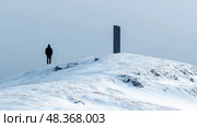 A person dressed in dark clothing stands alone on a snowy hill beside a tall monolith, surrounded by a bleak, gray sky. Стоковое AI-изображение, AI-художник Данила Большаков / Фотобанк Лори