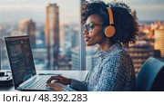 A woman with curly hair wears headphones while focusing on her computer screen in a sleek office. The sunset casts warm light over the cityscape, creating a productive atmosphere. Стоковое AI-видео, аниматор Данила Большаков / Фотобанк Лори