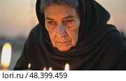 An elderly woman in a black chador lighting candles at dusk, their flickering glow reflecting in her grief-stricken eyes, surrounded by quiet reverence. Стоковое AI-видео, аниматор Sergey Grishin / Фотобанк Лори
