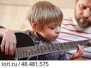 Handsome dad teaches his cute son to play the guitar sitting on sofa in the living room at home. Happy Father's Day. Father teaching his child to play guitar. Kid Learning music skill. Art education. Стоковое фото, фотограф Марина Демидюк / Фотобанк Лори