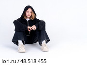 A caucasian teenage girl in a black hoodie with a hood is sitting on the floor on a white background. Studio photo. Difficult adolescence transitional age concept. Стоковое фото, фотограф Луиза Налимова / Фотобанк Лори