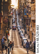 VALLETTA, MALTA - NOVEMBER 12, 2018 - Hilly ancient streets view with colourful balconies, people and cars in the medieval city. Редакционное фото, фотограф Сергей Богомяко / Фотобанк Лори
