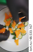 Professional chef wearing black gloves carefully adding chopped orange bell peppers into white bowl with sliced cucumbers on green cutting board during meal preparation. Стоковое видео, видеограф Данил Руденко / Фотобанк Лори