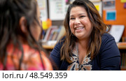 A woman engages in a conversation with a young girl inside a classroom setting. Стоковое AI-изображение, AI-художник Данила Большаков / Фотобанк Лори
