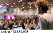 Young speaker addresses a large audience at a conference in a modern venue, showcasing public speaking skills and engagement. Стоковое AI-изображение, AI-художник Сергей Толмачёв / Фотобанк Лори