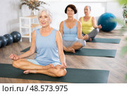 Elderly woman in company of hatha yoga lovers performs half-lotus pose, Ardha Padmasana. Стоковое фото, фотограф Яков Филимонов / Фотобанк Лори