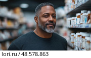 A man with a graying beard smiles while standing in a pharmacy aisle surrounded by shelves of medicine bottles. Стоковое AI-изображение, AI-художник Данила Большаков / Фотобанк Лори
