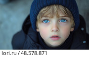 A young child with bright blue eyes looks up inquisitively, dressed warmly in a knitted hat against a cold backdrop. Стоковое AI-изображение, AI-художник Данила Большаков / Фотобанк Лори