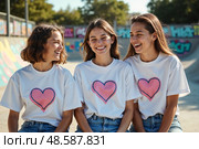 Три счастливые девочки подростка в одинаковых футболках с сердечками смеются вместе на улице. Стоковое AI-изображение, AI-художник Елена Болотина / Фотобанк Лори