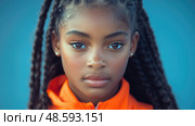 A young girl with intricate braids showcases her determination while wearing an orange jacket, framed by a vibrant blue backdrop. Стоковое AI-изображение, AI-художник Данила Большаков / Фотобанк Лори