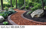 The curved path made of mulch traverses through vibrant plants and rocks in a peaceful garden setting. Стоковое AI-изображение, AI-художник Данила Большаков / Фотобанк Лори