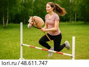 Hobby horsing. Energetic teenage girl jumping over hurdle with hobby horse toy during outdoor hobbyhorsing training on sunny summer day park. Стоковое AI-изображение, AI-художник Павел Муравьев / Фотобанк Лори