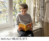 На школьном окне сидит мальчик с книгой в руках. Он внимательно читает, погружённый в свои мысли. За окном видны зелёные деревья, наполняющие атмосферу спокойствием. Стоковое AI-изображение, AI-художник Сергей Никифоров-Гордеев / Фотобанк Лори