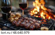 A cozy outdoor gathering features grilled meat and roasted potatoes by a crackling campfire, complemented by glasses of red wine. Стоковое AI-изображение, AI-художник Данила Большаков / Фотобанк Лори