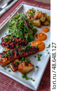 Grilled octopus with potatoes and greens on a white plate. Стоковое AI-изображение, AI-художник Olga Didraga / Фотобанк Лори