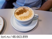 A creamy latte topped with decorative foam and crushed biscuits is presented on a saucer at a cozy cafe during midday hours. Стоковое фото, фотограф Данила Большаков / Фотобанк Лори