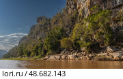 A calm red-brown river. There is green vegetation on the steep banks. Eroded limestone boulders near the water. Blue sky, clouds. Madagascar. Manambolo river. (2023 год). Стоковое фото, фотограф Вера Ивановна Кубарева / Фотобанк Лори