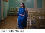 Woman in a blue dress holds a flute, sitting in a classical room with ornate chairs. The serene expression suggests contemplation or anticipation for a musical performance. Стоковое фото, фотограф Андрей Журавлев / Фотобанк Лори
