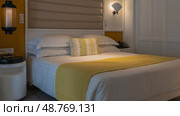 A double bed with white sheets and a yellow bedspread. Pillows at the headboard. The wall sconces are lit. Round bedside table. Louvered partition. Стоковое фото, фотограф Вера Ивановна Кубарева / Фотобанк Лори