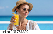 Tourist woman wearing sunglasses and sunhat drinking orange juice by the sea. Стоковое AI-изображение, AI-художник Юрий Поздников / Фотобанк Лори