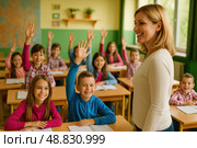 Учительница начальных классов улыбается и смотрит на учеников, активно участвующих в занятии. Дети сидят за партами в ярком, светлом классе, многие поднимают руки, желая ответить. На переднем плане — мальчик и девочка с тетрадями, на заднем — остальные ученики, сосредоточенные и вовлечённые. Класс оформлен зелёными и жёлтыми стенами, есть карта и растения. Сцена подчёркивает роль учителя в образовательном процессе, атмосферу доверия и взаимодействия между педагогом и учениками. Стоковое AI-изображение, AI-художник Sergey Starostin / Фотобанк Лори