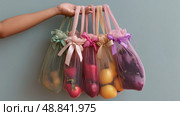 Hand holds colorful reusable mesh bags filled with various fruits against a muted backdrop. Стоковое AI-изображение, AI-художник Сергей Толмачёв / Фотобанк Лори