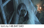 A woman dressed as a ghost peeks out from behind a window, her face partially obscured by a white veil. The image is dark and atmospheric, suggesting a spooky Halloween setting. Стоковое AI-изображение, AI-художник Данила Большаков / Фотобанк Лори