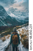 A lone female hiker, clad in winter gear, ascends a snowy mountain path with trekking poles. The path leads toward a towering mountain range with snow-capped peaks. Стоковое AI-изображение, AI-художник Данила Большаков / Фотобанк Лори
