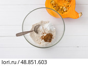 Glass bowl with oatmeal flour, baking powder and ground cinnamon, half a pumpkin with seeds on a white wooden background, top view. Stage of cooking delicious vegan dessert. Стоковое фото, фотограф Ольга Лепёшкина / Фотобанк Лори