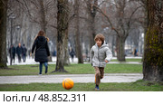 A homeless child playing with a ball in a public park, surrounded by winter trees, while adults walk by without noticing. Стоковое AI-изображение, AI-художник ALLA YURTAYEVA / Фотобанк Лори