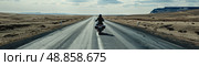 A lone motorcyclist rides down a straight, paved road through a barren, desert landscape. The sky is clear and blue, with a few fluffy clouds. Стоковое AI-изображение, AI-художник Данила Большаков / Фотобанк Лори