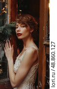 A woman in a white, beaded dress leans against a wooden doorway. She has short, wavy brown hair, bright red lipstick, and a delicate gold necklace. Стоковое AI-изображение, AI-художник Данила Большаков / Фотобанк Лори