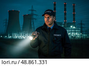 Security guard patrolling power plant at night, holding flashlight, wearing uniform, safety inspection, electric industry, night shift, infrastructure, security service, energy facility. Стоковое AI-изображение, AI-художник Павел Муравьев / Фотобанк Лори