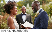 A joyous bride and groom exchange vows with an officiant smiling in the background, surrounded by greenery. Стоковое AI-изображение, AI-художник Данила Большаков / Фотобанк Лори
