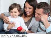 Toddler holding mobile telephone whilst sat with parents on sofa. Стоковое фото, агентство Ingram Publishing / Фотобанк Лори