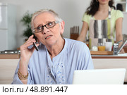 Elderly woman chatting on the phone whilst young helper works in the kitchen. Стоковое фото, агентство Ingram Publishing / Фотобанк Лори