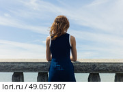 A young woman wearing a blue dress is standing by a wall with concrete balustrades on a promenade and is admiring the seaside on a sunny day. Стоковое фото, агентство Ingram Publishing / Фотобанк Лори