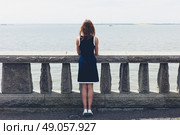 A young woman wearing a blue dress is standing by a wall with concrete balustrades on a promenade and is admiring the seaside on a sunny day. Стоковое фото, агентство Ingram Publishing / Фотобанк Лори