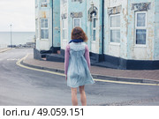 A young woman is standing in the street outside a blue house by the coast on a windy day. Стоковое фото, агентство Ingram Publishing / Фотобанк Лори