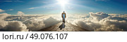 A solitary figure walks along a paved path, with a brilliant sun shining above. The individual is dwarfed by the vast expanse of clouds below. Стоковое AI-изображение, AI-художник Данила Большаков / Фотобанк Лори