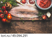 Raw fish fillet with frish herbs, tomatoes and sauce on dark rustic cutting board on dark aged wooden background, top view, border. Pollack fish cooking. Стоковое фото, агентство Ingram Publishing / Фотобанк Лори