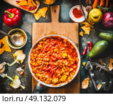 Vegetarian chili con carne dish in pan on wooden cutting board with spices and vegetables cooking ingredients on dark kitchen table background, top view. Mexican cuisine. Стоковое фото, агентство Ingram Publishing / Фотобанк Лори